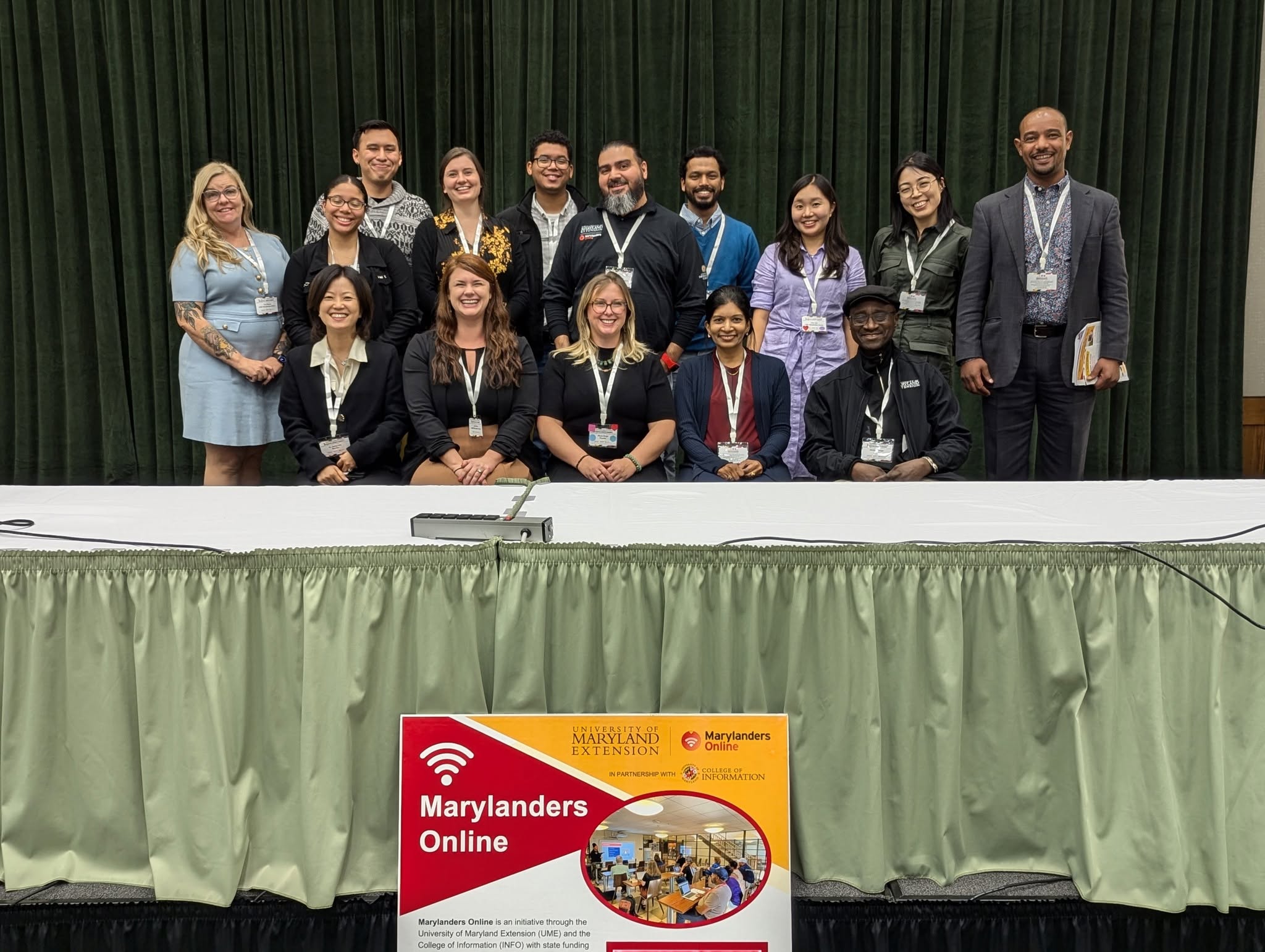 Two rows of UME Marylanders Online team members behind a green table with a UME Marylanders Online poster in the front center.