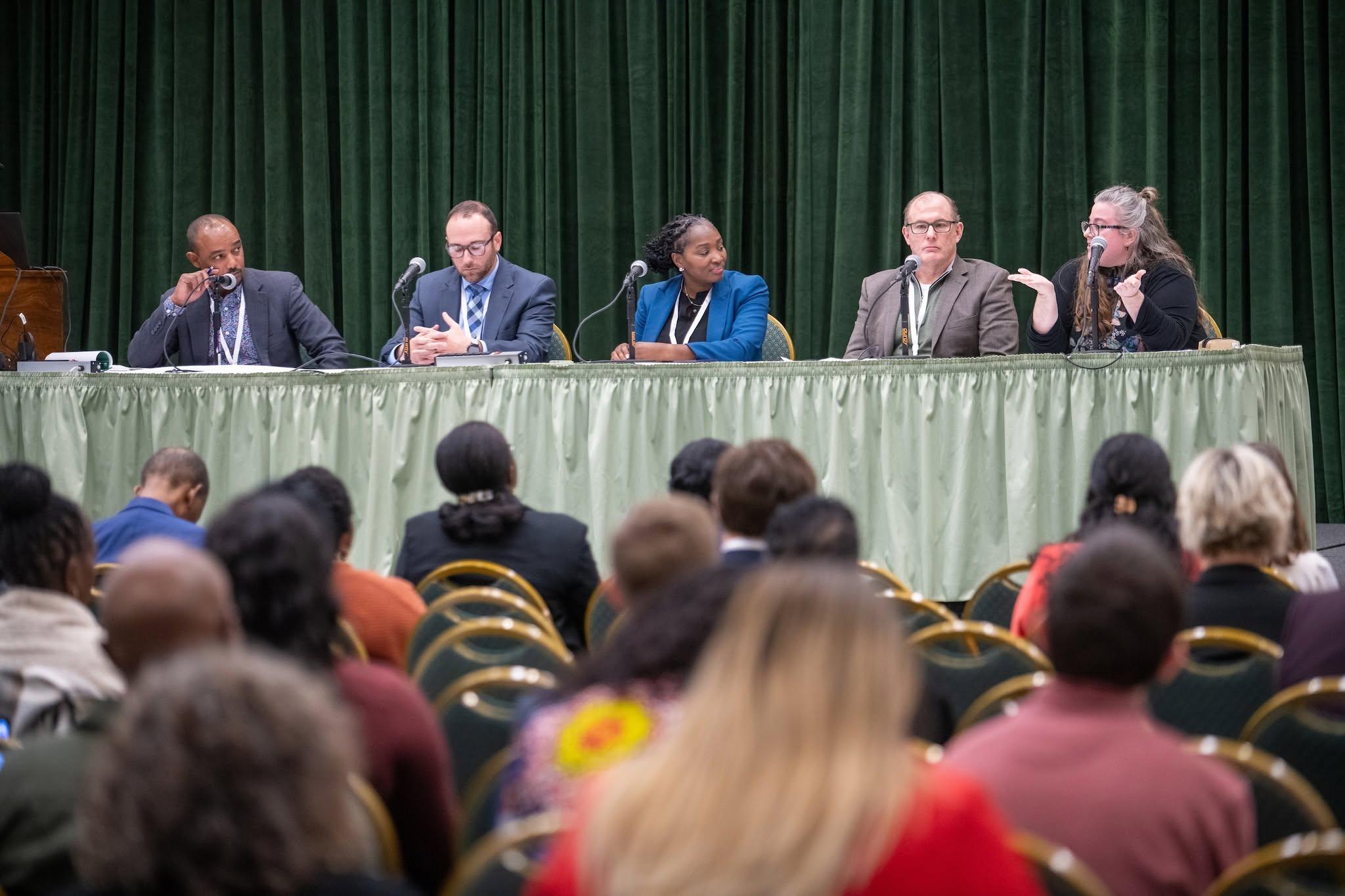 Picture from the 2024 Digital Opportunity Summit, featuring back of attendees heads facing a table of five panelists.