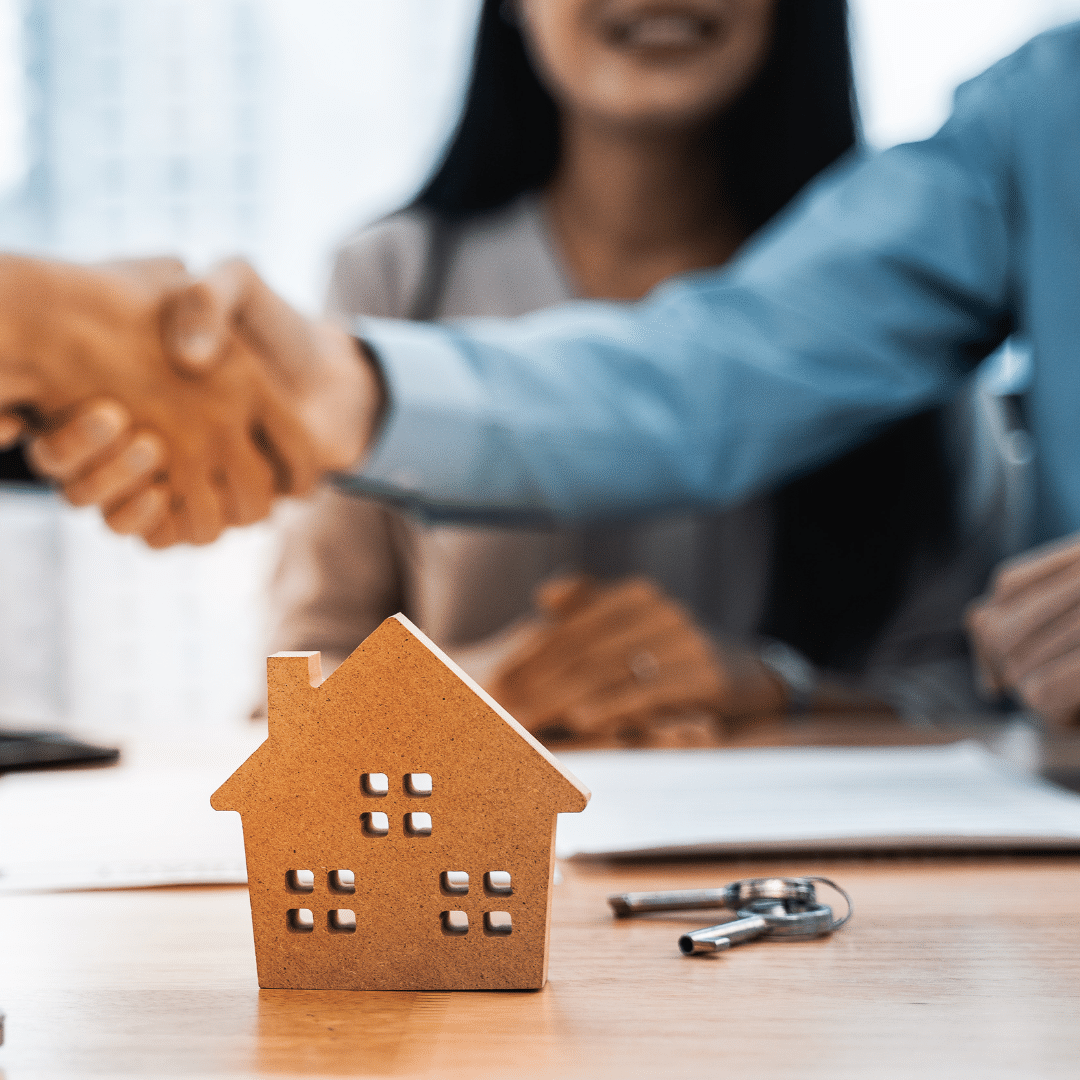 Graphic of a small gray model house placed on paperwork with a key, and people shaking hands above it.